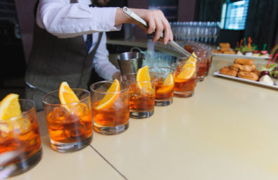 Bar Tavern Product Listing Image Bartender, barman preparing an alcoholic beverage, drink in nightclub, pub or bar. Man lays in a glass of whiskey with ice and slices of orange. glasses of whiskey or rum lined up.
