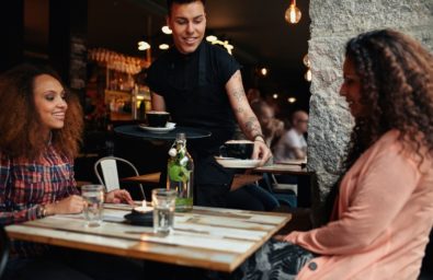 Waiter Serving Coffee To Young Women At Cafe Waiter serving coffee to young women at restaurant. Two female friends at coffee shop.